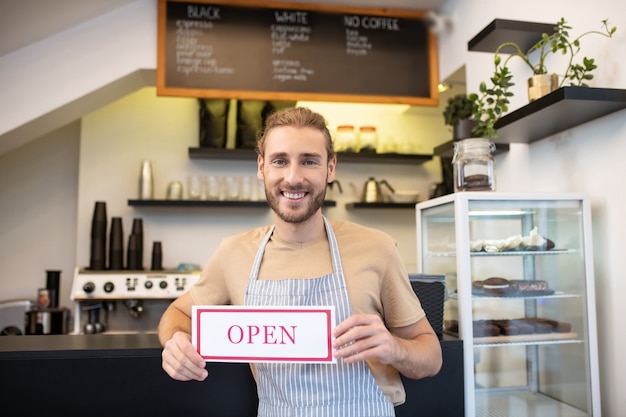 Invitation, café. Homme satisfait en t-shirt et tablier debout près du comptoir dans son café d'ouverture invitant les visiteurs