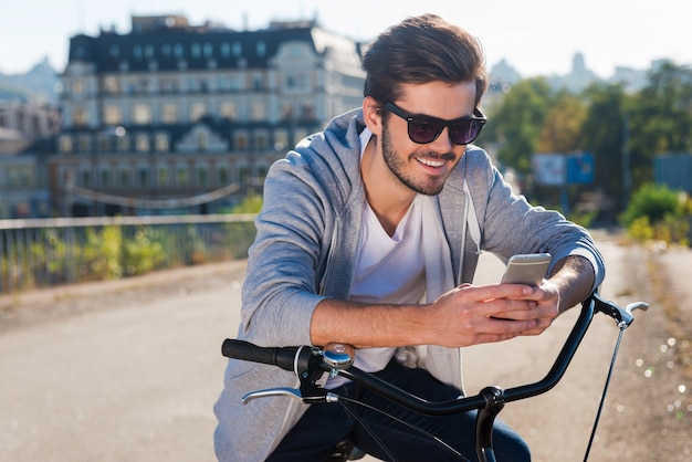 Inviter un ami pour un tour. Beau jeune homme souriant se penchant sur son vélo et regardant son téléphone portable tout en se tenant à l'extérieur