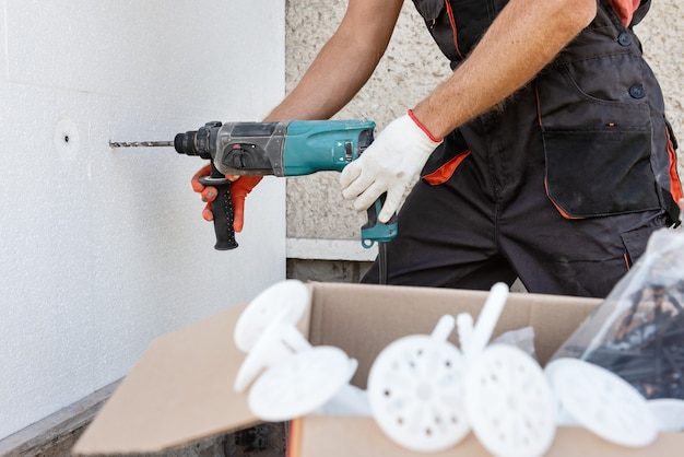 Isolation de la maison avec polyfoam. Le travailleur perce des trous pour fixer le panneau de polystyrène sur la façade.