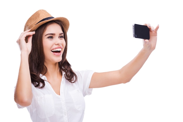 J'adore les selfies ! Cheerful young woman in funky hat faisant selfie avec son téléphone intelligent et souriant en se tenant debout sur fond blanc