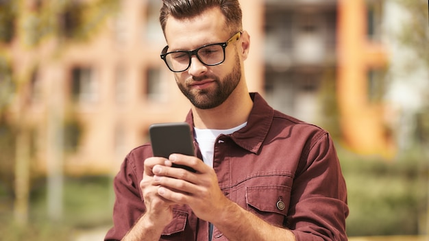 J'ai besoin d'appeler. Bel homme barbu en tenue décontractée et lunettes regardant son smartphone en se tenant debout dans la rue. Mode de vie. Numérique. la communication