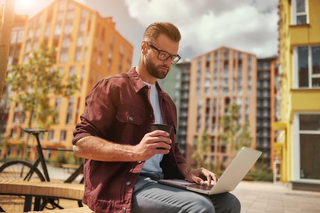 J'ai besoin de café bel homme avec du chaume dans des vêtements décontractés et des lunettes travaillant sur un ordinateur portable