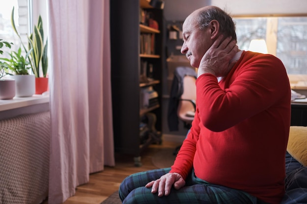 J'ai besoin d'un massage. Homme senior frustré en pull rouge tenant la main sur son cou assis sur un canapé