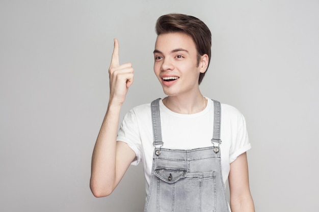 J'ai une idée. Portrait d'un jeune homme brune surpris dans un style décontracté avec un t-shirt blanc et une salopette en jean debout et regardant ailleurs et a eu l'idée. tourné en studio intérieur, isolé sur fond gris.