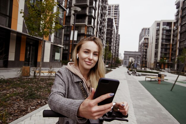 J'aime beaucoup cette application de location de scooters! Jolie femme en manteau s'appuie sur le volant d'un scooter, tient un smartphone dans sa main et regarde la caméra.