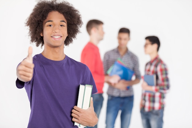 J'aime étudier! Joyeux adolescent africain tenant des livres et faisant des gestes pendant que ses amis se tiennent sur l'arrière-plan