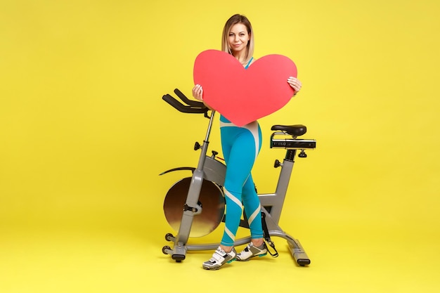 J'aime le sport Portrait complet d'une femme assise sur un canapé de fitness debout près d'un vélo d'exercice et tenant un grand coeur rouge portant des vêtements de sport bleus Prise de vue en studio intérieure isolée sur fond jaune