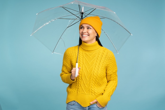 J'aime un temps différent. Jeune femme brune portant un bonnet et un pull tenant un parapluie transparent isolé sur un mur bleu avec une expression heureuse