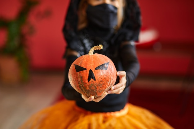 Jack o lantern, citrouille d'halloween en masque noir de protection médicale dans les mains de la fille lors de la célébration d'Halloween à la maison pandémie de coronavirus Covid19, espace de copie