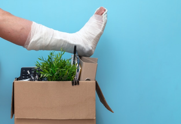 Jambe en plâtre sur une boîte d'un homme viré du travail sur un fond bleu