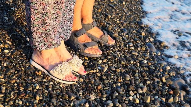 Les jambes du couple en sandales se tiennent côte à côte sur une plage de galets au coucher du soleil