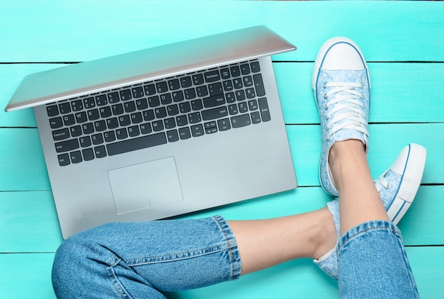 Jambes féminines en jeans et baskets, ordinateur portable sur un plancher en bois turquoise. Technologies modernes. Génération z. Vue de dessus. Mise à plat.