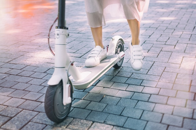 Jambes de femme sur scooter électrique de transport écologique sur carrelage de trottoir en journée ensoleillée d'été