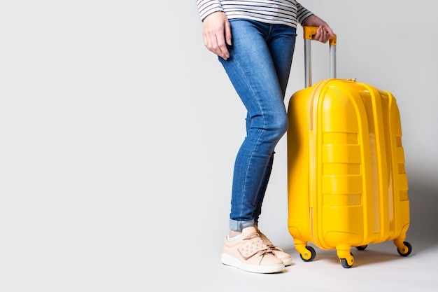 Les jambes des femmes en jeans se tiennent à côté d'une valise jaune sur fond clair