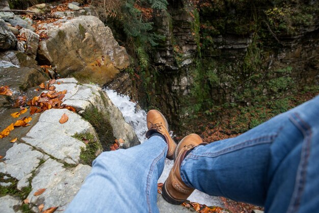 Jambes d'homme en jeans et bottes marron assis sur le bord regardant la cascade