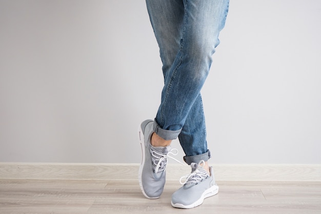 Jambes d'un jeune homme en jeans et baskets sur un mur gris.