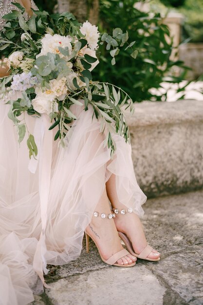 Photo les jambes de la mariée assises près de l'arbre dans la vieille ville avec un bouquet de roses blanches, de pivoines et de branches d'eucalyptus en gros plan