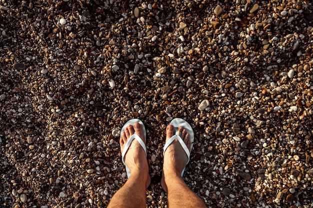 Jambes masculines en chaussons de plage, sur une robe de galets.