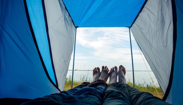 Jambes nues de l'homme et de la femme tendues hors de la tente touristique. Vue sur la rive du fleuve avec herbe verte et ciel bleu aux beaux jours d'été. Traversée pieds nus d'amoureux se touchant. Tourisme familial, lune de miel.