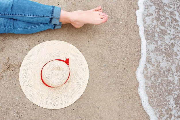 jambes et pieds d'une femme caucasienne en jeans assise au bord de la mer sur la côte avec un chapeau à proximité sur le sable