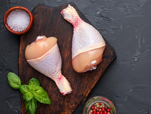 Jambes de poulet crues avec des épices sur une planche à couper en bois sur un fond noir en ardoise ou en béton