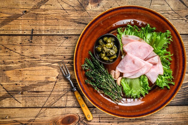 Jambon fumé tranché avec salade fraîche et câpres sur une assiette. fond en bois. Vue de dessus. Espace de copie.