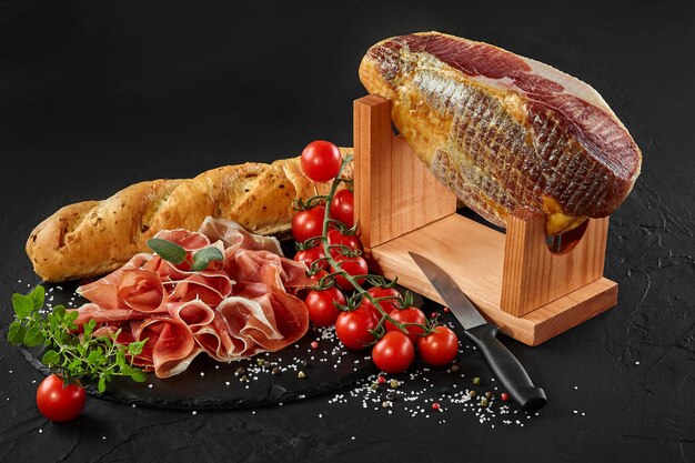 Jamon sculpté de la jambe de porc sur le stand de jamonera tomates cerises herbes sur ardoise en pierre noire baguette et couteau sur fond sombre Gros plan