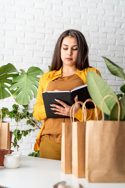 Jardinage domestique. Petite entreprise. Jeune femme jardinier prendre des notes dans le bloc-notes, ligne de sacs à provisions artisanales pour l'usine d'emballage à l'avant