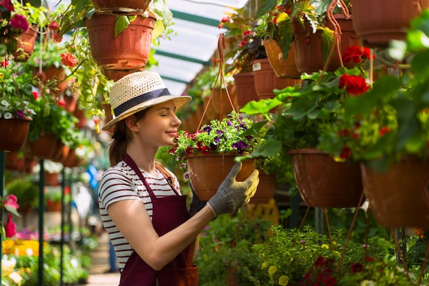 Le jardinier de femme dans le chapeau et les gants travaille avec des fleurs dans la serre chaude
