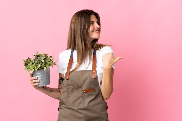 Jardinier femme tenant une plante isolée sur fond rose pointant vers le côté pour présenter un produit