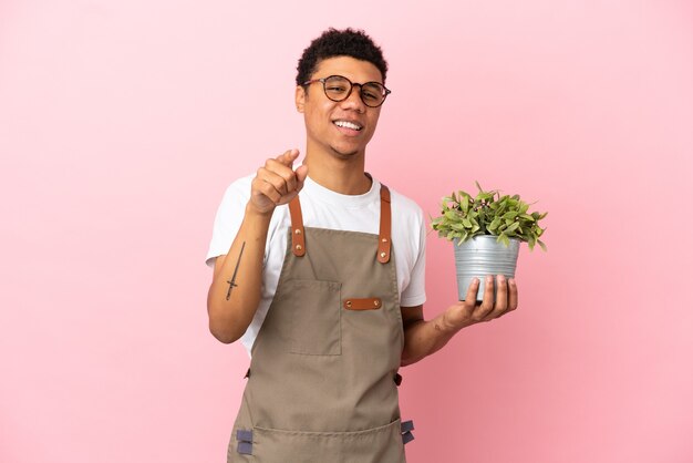 Jardinier homme africain tenant une plante isolée sur fond rose surpris et pointant vers l'avant