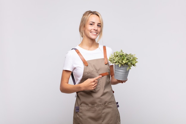 Jardinier de jeune femme souriant joyeusement, se sentant heureux et pointant vers le côté et vers le haut