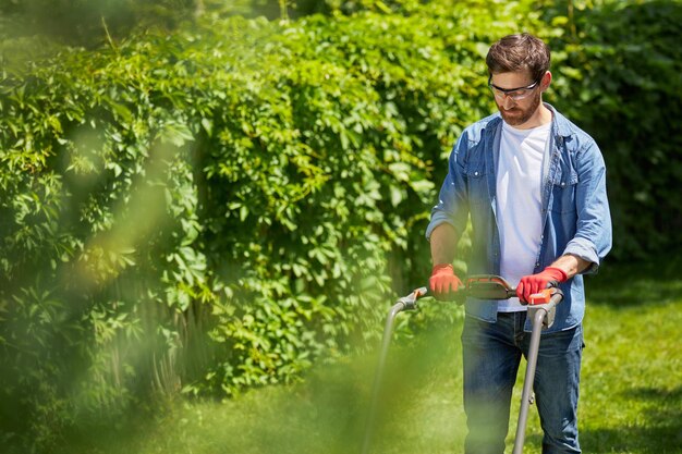 Photo jardinier masculin sérieux en gants poussant la poignée d'une tondeuse à gazon moderne tout en coupant l'herbe en été