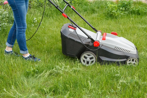 Une jardinière en jeans fonctionne avec une tondeuse à gazon dans le jardin en été. Équipement de tondeuse à gazon. Outil de travail de soin de jardinier de tonte.