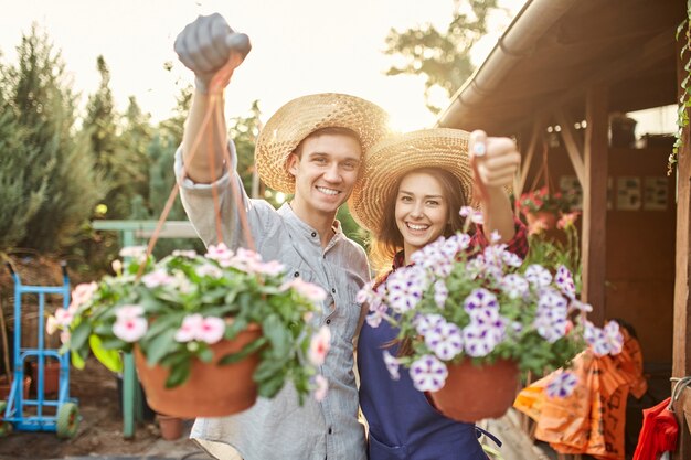 Les jardiniers heureux de type et de fille dans des chapeaux de paille tiennent des pots avec le pétunia merveilleux dans le jardin sur un soleil.