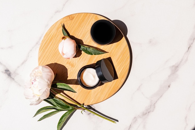 Des jarres ouvertes et fermées avec de la crème pour soigner la peau du visage et du corps sur un podium rond en bois avec des péonies vivantes Cosmétiques naturels Top view