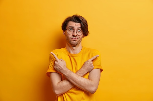 Je me demande quoi choisir. Un hipster hésitant croise les mains sur la poitrine dans différentes directions vêtu d'un t-shirt décontracté isolé sur un mur jaune sans savoir quelle est la meilleure décision