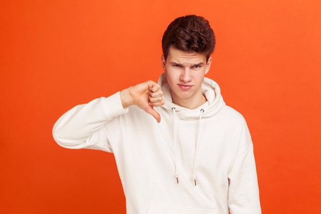 Je n'aime pas ce jeune homme en sweat à capuche de style décontracté montrant le signe du pouce vers le bas avec dégoût sur son visage test d'application retour tourné en studio intérieur isolé sur fond orange