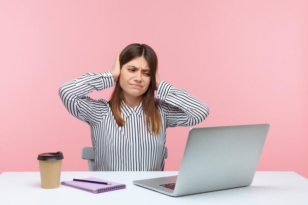 Je ne t'entends pas. Une employée de bureau bouleversée couvrant les oreilles et regardant avec irritation l'écran de l'ordinateur portable, agacée par un son désagréable pendant l'appel vidéo. Studio intérieur tourné isolé sur fond rose