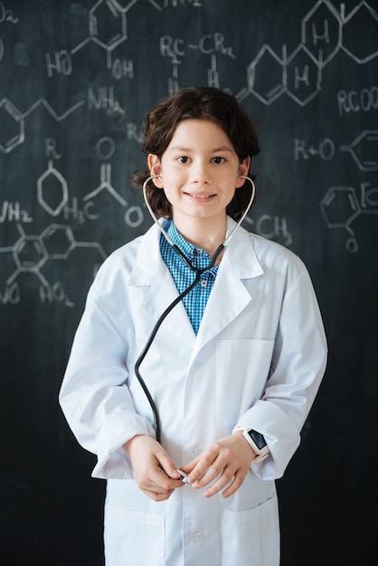 Je pense à ma future carrière. Intelligent souriant joyeux garçon debout près du tableau noir à l'école tout en ayant une leçon de médecine et à l'aide d'un stéthoscope