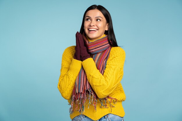 Je souhaite que. Femme brune souriante portant un pull jaune tricoté et une écharpe tenant ses mains près de sa tête et rêvant au studio sur fond bleu