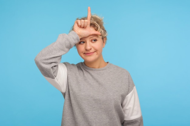 Je suis perdant Femme frustrée aux cheveux courts et bouclés en sweat-shirt montrant un geste boiteux ou perdant avec les doigts L sur le front contrarié par l'échec de la perte d'emploi tourné en studio intérieur isolé sur fond bleu