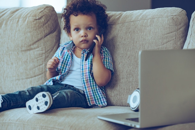 Photo je suis tellement occupé ! petit garçon africain parlant au téléphone portable et regardant la caméra alors qu'il était assis sur le canapé à la maison