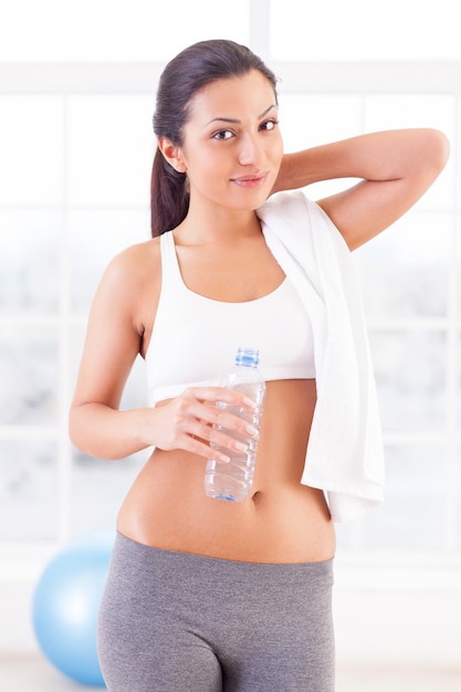 Je suis toujours en forme. Jolie jeune femme en vêtements de sport tenant une bouteille d'eau et regardant la caméra en se tenant debout dans un club de sport