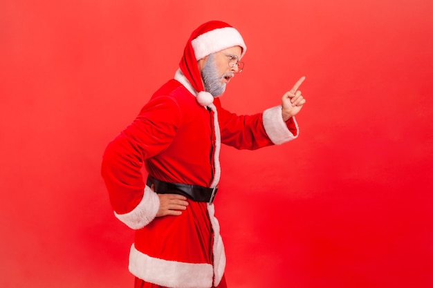 Je t'ai dit! Vue latérale d'un homme âgé à la barbe grise portant un costume de père Noël debout avec un signe d'attention, avec un geste d'avertissement du doigt. Studio intérieur tourné isolé sur fond rouge.