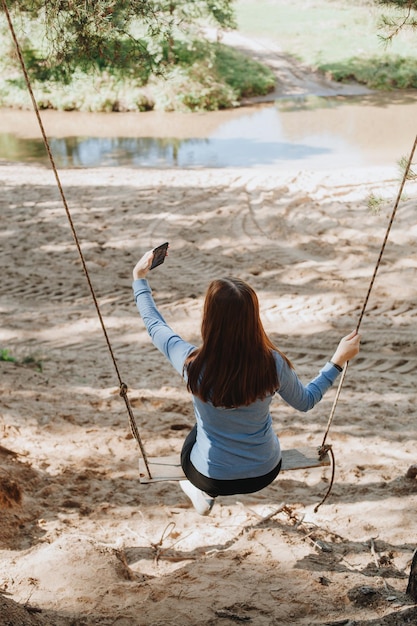 Je veux me souvenir de ce jour. Portrait d'une charmante fille prenant une photo avec un smartphone assis sur une balançoire en bois. Elle sourit