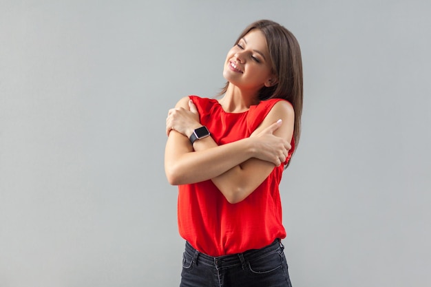 Je vis moi-même. Portrait d'heureuse belle jeune femme brune en chemise rouge debout, sourire à pleines dents et se serrant dans ses bras et s'amusant. intérieur, tourné en studio, isolé sur fond gris.