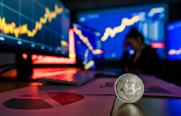 Jeton de crypto-monnaie bitcoin doré sur le bureau devant le graphique d'analyse financière rapport de croissance des échanges sur l'écran de l'ordinateur et de l'ordinateur portable pendant que le courtier se réunit en arrière-plan flou d'ombre.