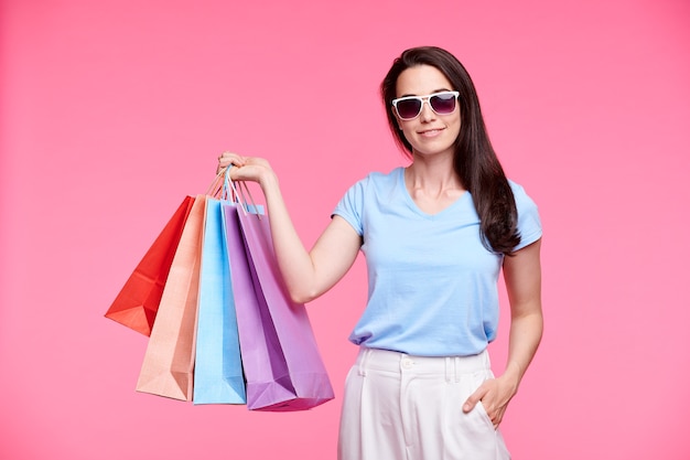 Jeune accro du shopping élégant dans des lunettes de soleil et des vêtements décontractés se vantant de ses achats tout en tenant un tas de sacs en papier sur un mur rose