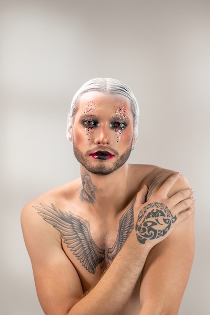 Jeune acteur de théâtre moderne avec maquillage de scène, cheveux blancs dans une coiffure précise et tatouage sur la poitrine, le cou et la main vous regardant isolément